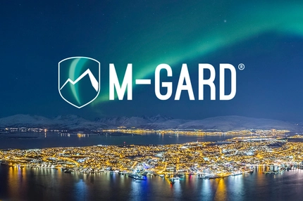 Image M-GARD logo over a brightly lit Arctic coastal city beneath green northern lights and snow-covered mountains.