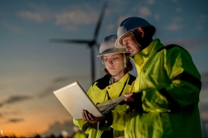Image Two people in high vis clothes inspect paperwork next to windmills