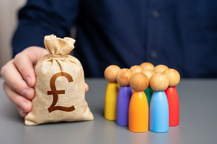 Image A businessman holds a british pound sterling money bag near a group of people figurines.