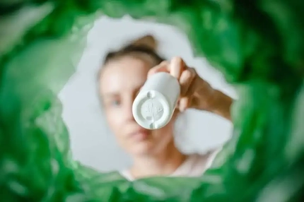 Image Woman holding a white bottle toward the camera, framed by lush green leaves in a soft-focus background.