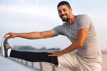 Image Smiling man wearing earphones stretches one leg outdoors while dressed in athletic clothing.