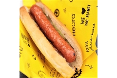 Image Plant-based hot dog in a bun served on branded yellow Heura wrapper with sustainability messaging.