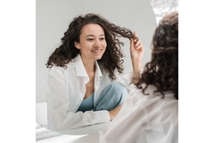 Woman with curly hair plays with her hair while smiling at her reflection in a mirror.