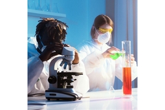 Image One scientist looks into a microscope while another pours a green chemical from an erlenmeyer glass flask into a beaker.