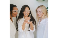 A Black woman, Asian woman and Caucasian woman wearing lab goggles observe a leaf contained in a test tube.