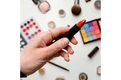 A woman's hand holding up a lipstick, while in the background there is a variety of cosmetic products scattered across a table surface.
