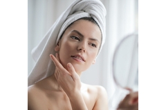 Image A woman with her hair wrapped in a towel applies face cream while looking into a mirror.