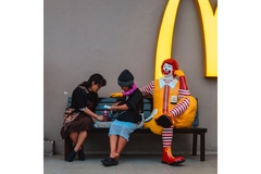 Image Two women sitting on a bench eating McDonalds with a Ronald McDonald statue sitting on the bench.