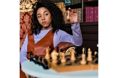 Image A woman sits in front of table with a chessboard on it in a library and holds the queen up so her competitor can see it.