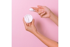 Image against a plan background one hand holds up an open cream, the other hand hover above it with index and middle fingers covered in cream