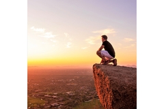 Image A man kneels on a cliff edge as the sun sets.