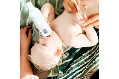 Image Baby boy laying holding a plastic bottle to his mouth while being applied with moisturizer on belly.