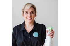 smiling woman in cleaners uniform with a badge on her breast holds a cleaning spray in her right hand.