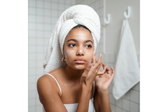 Image A woman looking in the mirror with a towel on her head while applying face cream.