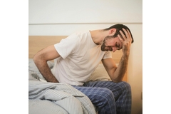 Image A man sits on the edge of his bed and holds his head and stomache as if in pain.