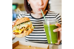 Image A woman holding a vegan burger and sipping green smoothie with a straw.