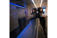 A woman stands next to a bunch of servers while checking her laptop.