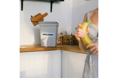woman with a banana in the foreground with a white grey bin in the background on the kitchen counter labeled organic