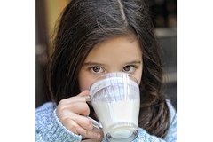 Image A young girl in blue sweater sipping milk from a transparent cup.