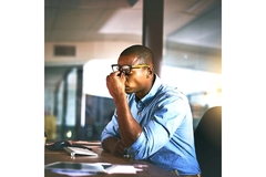 Image A young man sits in front of a computer at his office, pinching between his eyes like he is feeling some stress.