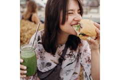 Image A young girl biting into a vegetarian burger while holding a green smoothie in her right hand.