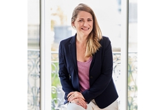 Image A woman in a navy blue coat and pink top sitting with one hand over the other and smiling.