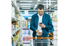Image A young man in a denim jacket looking at his phone in a supermarket.