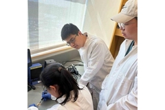 Image Three people in lab coats, look at one of them work on the patch on a table