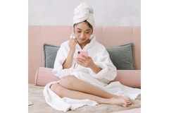 woman in bath robe on bed with undereye patch looks at phone