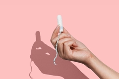 Image A hand holding a tampon in front of a pink background.