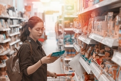 Image a young woman holding a shopping cart and looking at a food packet.