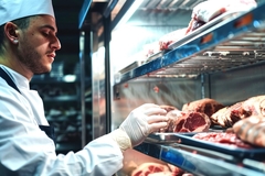 Image Butcher arranging trays of fresh meat cuts on racks inside a walk-in freezer.