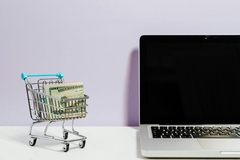 Image A laptop next to a mini shopping cart with cash in it.