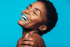 A woman with yellow eyeliner and blue lipstick and smiling in front of a blue background.