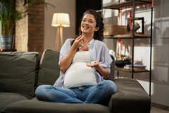 A pregnant women smiles while sitting on the couch and preparing to take a supplement.