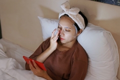 Image Woman laying in bed wearing sheet mask on her face and watching her phone