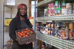 Image Black woman holds a basket of tomatos in next to shelves of canned food