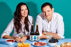 Image a man and a woman in white shirts sitting behind a blue table with two bottles of olive oil and berries against a green background