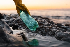 A hand in a glove holding a plastic bottle on beach.
