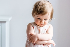A sad looking toddler crosses her arms and looks to the ground.