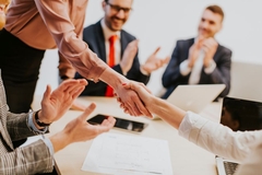 Image A group of people in a bright business office applaud as two women shake hands on a business deal.