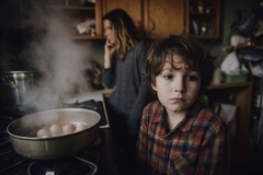 Image A young boy stands somberly in the kitchen as breakfast is being prepared with a pensive expression.