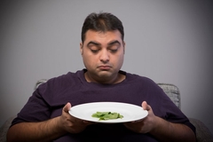 Image An overweight man stares sadly at a couple of peices of lettuce on the plate he is holding at the dinner table.