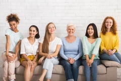 Image Several happy women of all ages and demographics sitting on a couch and laughing.