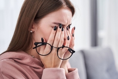 Image A woman holds her glasses in her hand and rubs her eyes while sitting on a white couch.