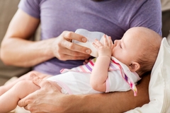 A man wearing a perriwinkle colored shirt bottle feeds a beautiful baby while sitting on the couch.