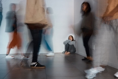 Image A girl sitting in a corner while people walk around her.