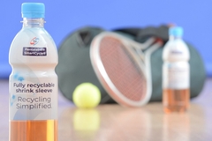 Plastic bottle with full-body shrink sleeve label promoting recyclability, placed in a fitness setting with tennis gear in the background