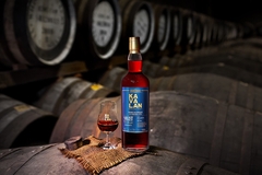 A bottle of Kavalan Solist Vinho Barrique Single Cask Strength whisky and a filled tasting glass resting on a barrel in a dimly lit aging cellar lined with oak casks.