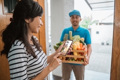 Image a woman holding a smartphone and a delivery man with a crate of fruits and vegetables at the door
